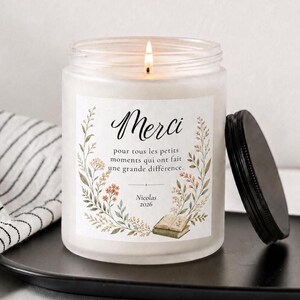May include: A frosted glass candle in a jar with a black lid, featuring a label with the text "Merci pour tous les petits moments qui ont fait une grande différence. Nicolas 2026" surrounded by a floral wreath and an illustration of books.