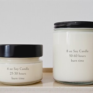 May include: Two soy wax candles in glass jars with black lids. The smaller candle is labeled "4 oz Soy Candle" with a 25-30 hour burn time. The larger candle is labeled "8 oz Soy Candle" with a 50-60 hour burn time.