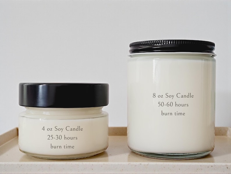 May include: Two soy wax candles in glass jars with black lids. The smaller candle is labeled "4 oz Soy Candle" with a 25-30 hour burn time. The larger candle is labeled "8 oz Soy Candle" with a 50-60 hour burn time.