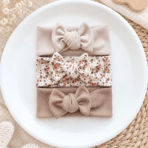 Könnte beinhalten: Ein Set aus drei gerippten Stirnbändern mit geknoteten Schleifen. Das obere Stirnband ist einfarbig beige, das mittlere zeigt ein kleines Blumenmuster mit orangefarbenen und braunen Blüten auf cremefarbenem Grund, das untere ist einfarbig hellbraun.