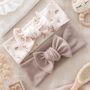 Könnte beinhalten: Zwei Stoffstirnbänder mit Knoten-Schleifen. Ein Stirnband ist cremefarben mit braunem Blumenmuster, das andere ist einfarbig taupe. Das Bild enthält Symbole mit den Beschriftungen: Perfect for babies & toddlers, Made with Oeko-Tex certified cotton, Stretchy for a perfect fit, und Handmade with love.