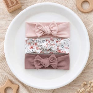 Könnte beinhalten: Drei Stirnbänder mit geknoteten Schleifen, die vertikal angeordnet sind. Das obere Stirnband ist einfarbig hellrosa, das mittlere hat einen weißen Hintergrund mit einem Blumenmuster in Rosa- und Grüntönen, das untere ist einfarbig altrosa.