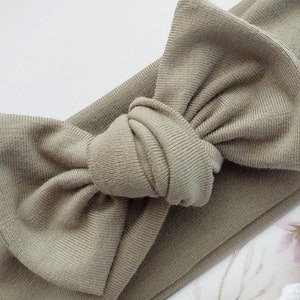 May include: A sage green knotted headband with a large bow. The headband is made of a soft, stretchy fabric.