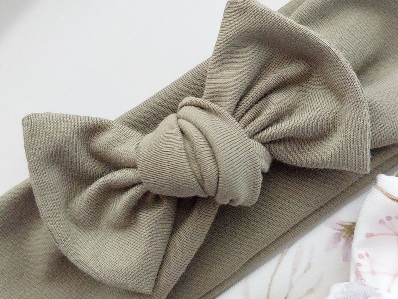 May include: A sage green knotted headband with a large bow. The headband is made of a soft, stretchy fabric.