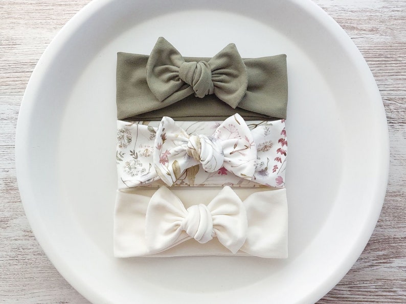 May include: Three fabric headbands with knotted bows. One headband is solid olive green, one is white with a floral print, and one is solid off-white.