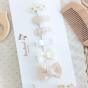 Peut inclure: Un ensemble de six barrettes à cheveux sur un support blanc marqué Bowland Accessories. La collection comprend une barrette marguerite blanche, deux barrettes ovales en tissu beige à pois et vichy, une barrette ruban blanc et deux nœuds en tissu beige.