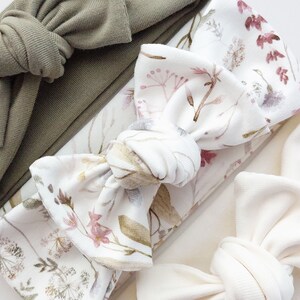 May include: Three fabric headbands with different patterns. The first headband is a solid sage green color. The second headband has a white background with a floral pattern in shades of pink, brown, and green. The third headband is a solid off-white color.