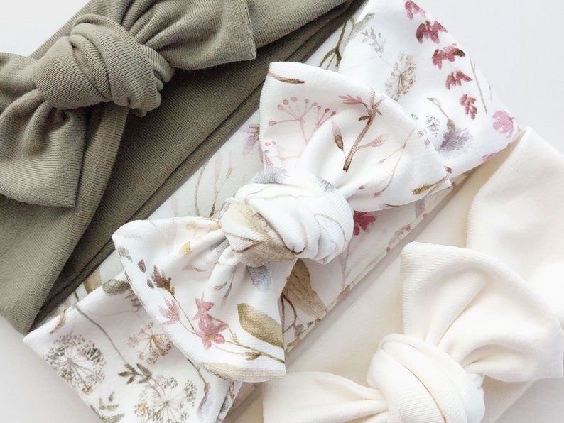 May include: Three fabric headbands with different patterns. The first headband is a solid sage green color. The second headband has a white background with a floral pattern in shades of pink, brown, and green. The third headband is a solid off-white color.
