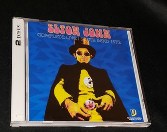 May include: A CD case with a blue and white background featuring Elton John in a yellow suit and black cape. The text on the case reads "Elton John Complete Live | South Bend 1972".