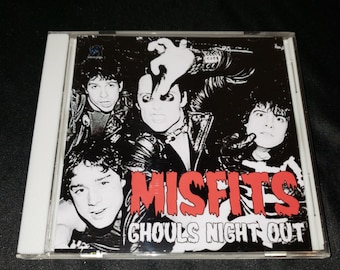 Może przedstawiać: Pudełko CD z zespołem Misfits. Okładka jest czarno-biała, z twarzami członków zespołu i czerwonym napisem "MISFITS" oraz białym "GHOULS NIGHT OUT".