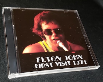 May include: A CD case featuring Elton John with sunglasses and a microphone. The album cover reads "ELTON JOHN FIRST VISIT 1971" in white text against a dark background. The CD is in a clear plastic case.