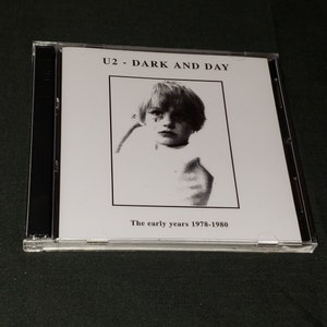 May include: A black and white CD case with a black and white photo of a young boy on the front. The text "U2 - DARK AND DAY" is printed in black at the top of the case. The text "The early years 1978-1980" is printed in black at the bottom of the case.