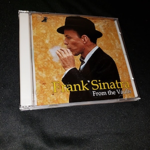 May include: A CD case with a photo of Frank Sinatra on the front. The photo shows Sinatra in a black suit and hat, smoking a cigarette. The text on the case reads "Frank Sinatra From the Vaults".