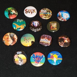May include: A collection of 15 round, colorful, button-style pins featuring the band Styx. The pins feature various album covers and logos.