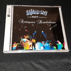 May include: A CD case with a black background and a photo of a band on stage. The text on the case reads "Genesis Live at the Roxy Theatre 1973 Antiques Roadshow".