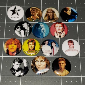 May include: A collection of 12 round button pins featuring images of David Bowie in various styles and outfits. The buttons are colorful and feature a variety of images, including Bowie in a suit, with a lightning bolt on his face, and with a red and white striped shirt.