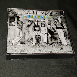 May include: Black and white photo of a band, Genesis, on a CD case. The band members are sitting on a tree trunk and a brick wall. The album title, "Lamb Sessions", is printed on the case.