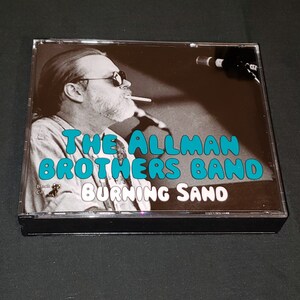 May include: A black and white photo of a man with a beard and sunglasses, holding a cigarette. The photo is on a CD case with the text "The Allman Brothers Band Burning Sand" in teal blue letters.