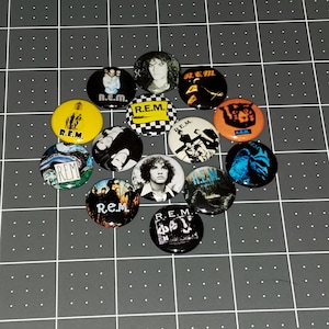 May include: A collection of 13 round buttons featuring various images of the band R.E.M. The buttons are black, white, yellow, orange, and blue with different images of the band members and album covers.