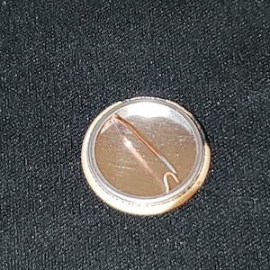 Puede incluir: La parte posterior de un bot&oacute;n redondo con un respaldo de metal plateado y un pasador dorado. El bot&oacute;n tiene un dise&ntilde;o en blanco y negro en la parte delantera.