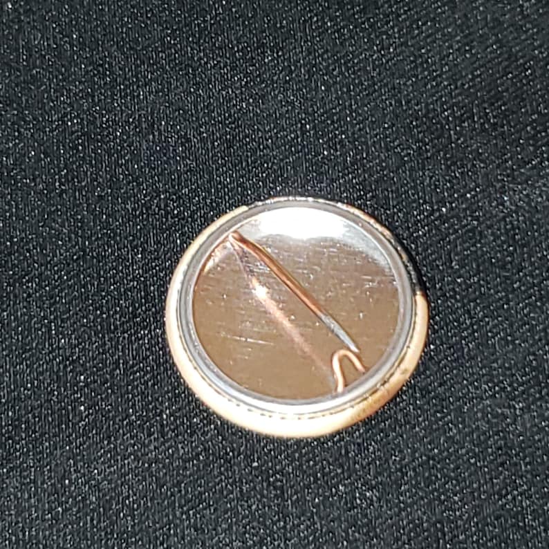Puede incluir: La parte posterior de un bot&oacute;n redondo con un respaldo de metal plateado y un pasador dorado. El bot&oacute;n tiene un dise&ntilde;o en blanco y negro en la parte delantera.