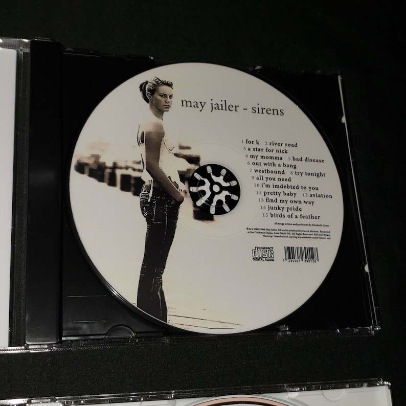 May include: A CD case with a white CD inside. The CD features a photo of a woman with a white shirt and blue jeans. The text "may jailer - sirens" is printed on the CD. The track list is printed on the CD, including the song titles "for k", "river road", "a star for nick", "my momma", "bad disease", "out with a bang", "westbound", "try tonight", "all you need", "i'm indebted to you", "pretty baby", "aviation", "find my own way", "junky pride", and "birds of a feather".