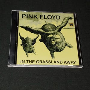May include: A yellow CD case with a black spine. The front of the case features a black and white illustration of two flying pigs against a yellow background. The text "PINK FLOYD" is printed in black at the top of the case and "IN THE GRASSLAND AWAY" is printed in black at the bottom of the case.