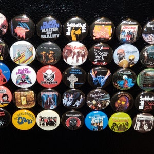 May include: A collection of 36 Black Sabbath album cover buttons. The buttons are round and feature various album covers from the band's discography. The buttons are arranged in a grid pattern on a black surface.