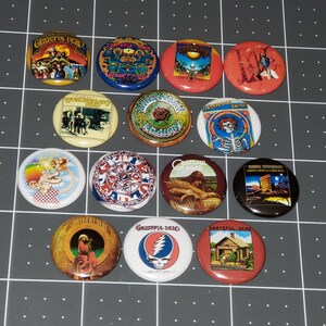 May include: A collection of 14 round Grateful Dead band pins. The pins feature various images, including the band's iconic skull and roses logo, a dancing bear, and a building. The pins are all in different colors and designs.