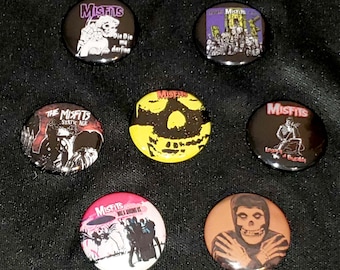 Puede incluir: Siete pines redondos de la banda Misfits. Los pines muestran diferentes portadas de álbumes y logotipos de la banda. Los pines son negros, amarillos, rojos y blancos.