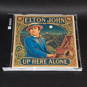May include: A CD case featuring Elton John's album "Up Here Alone." The cover art depicts Elton John in a blue suit and hat playing a piano, with a cityscape backdrop. The case is labeled "2 DISCS."