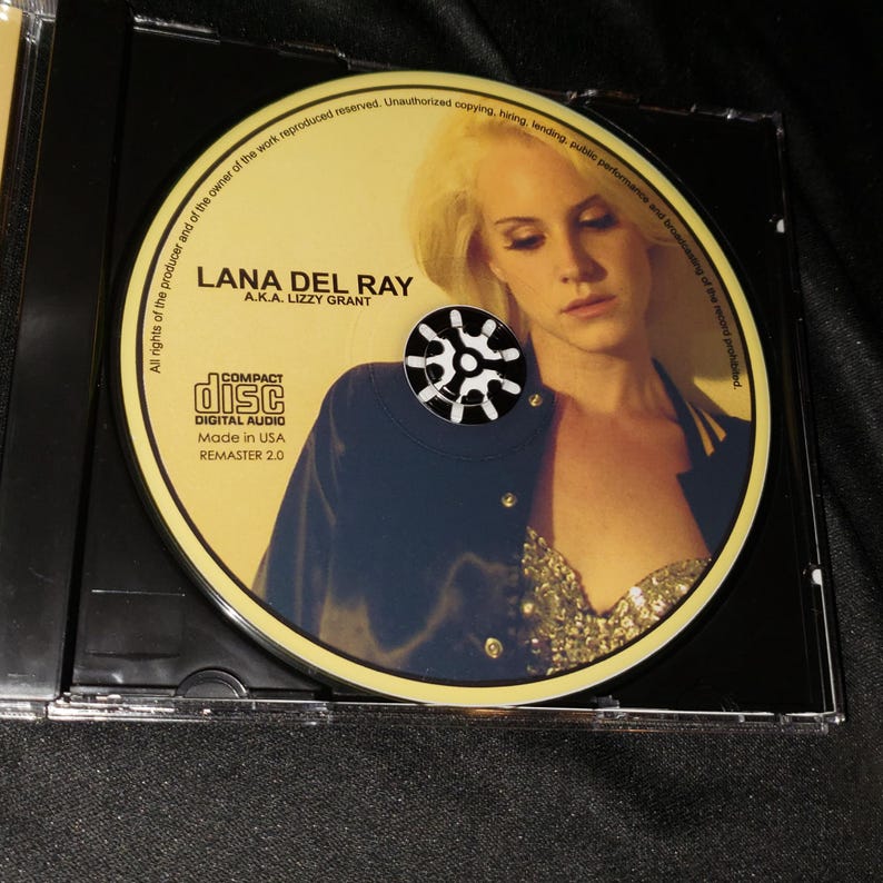 May include: A yellow CD with a black and white compact disc logo. The CD features a photo of Lana Del Ray, a.k.a. Lizzy Grant, in a black jacket and a sparkly top. The text "LANA DEL RAY" and "A.K.A. LIZZY GRANT" is printed in black on the CD. The text "COMPACT DISC DIGITAL AUDIO" is printed in black on the CD. The text "Made in USA" and "REMASTER 2.0" is printed in black on the CD.