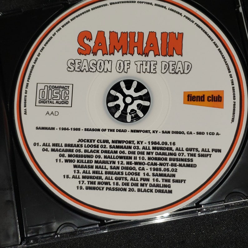 May include: A white CD with a black and white image of a spinning wheel and the text "SAMHAIN SEASON OF THE DEAD" in black. The CD is labeled with the band's name, album title, and track list. The text "fiend club" is printed in white on a black background.