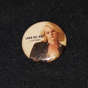 Puede incluir: Un bot&oacute;n redondo con una imagen de Lana Del Rey. El bot&oacute;n es beige con texto negro que dice "Lana Del Rey" y "...Lizzy Grant".