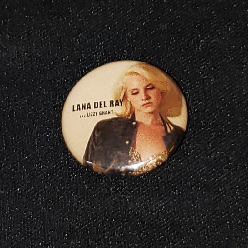 Puede incluir: Un bot&oacute;n redondo con una imagen de Lana Del Rey. El bot&oacute;n es beige con texto negro que dice "Lana Del Rey" y "...Lizzy Grant".