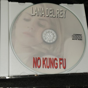 Puede incluir: Un CD con una imagen borrosa de la cara de una mujer en la parte delantera. El texto "Lana Del Rey" est&aacute; impreso en negro en la parte superior del CD. El texto "No Kung Fu" est&aacute; impreso en rojo en la parte inferior del CD. El CD est&aacute; en una caja de pl&aacute;stico transparente.