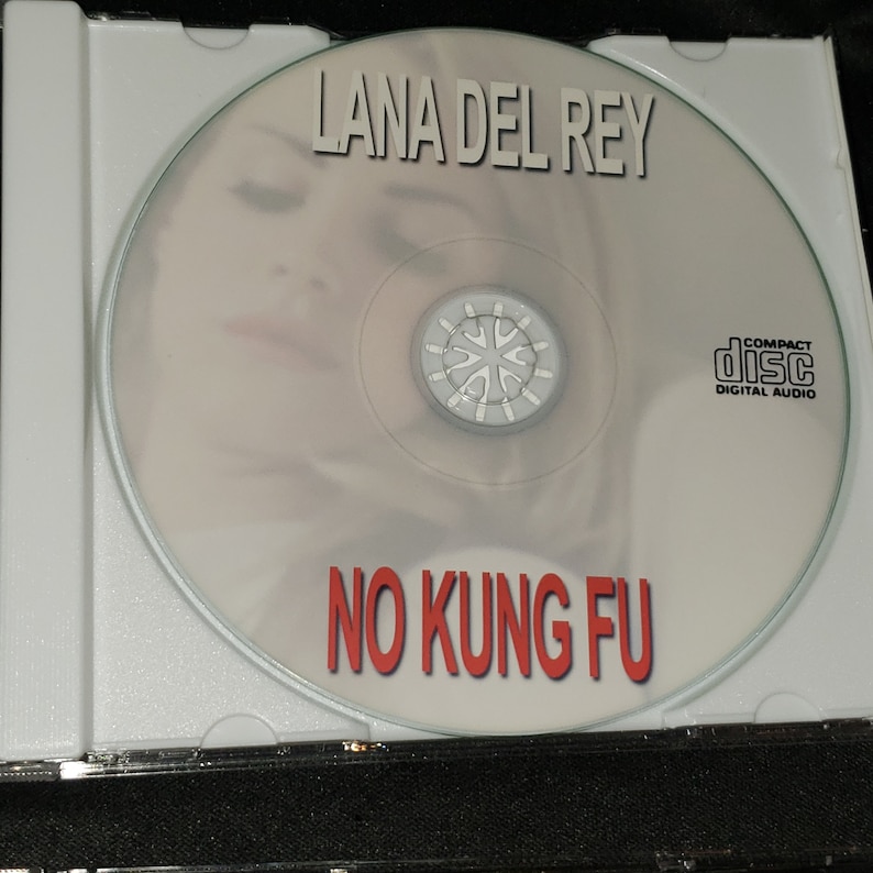 Puede incluir: Un CD con una imagen borrosa de la cara de una mujer en la parte delantera. El texto "Lana Del Rey" est&aacute; impreso en negro en la parte superior del CD. El texto "No Kung Fu" est&aacute; impreso en rojo en la parte inferior del CD. El CD est&aacute; en una caja de pl&aacute;stico transparente.