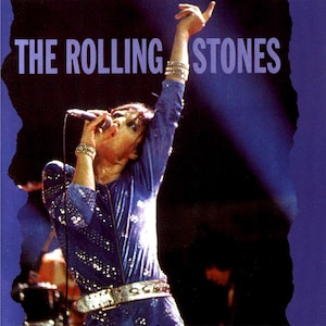 Puede incluir: Imagen de una actuación en vivo de The Rolling Stones. El cantante, con un mono azul con lentejuelas y cinturón plateado, sostiene un micrófono y levanta un brazo. El nombre de la banda aparece en texto morado en la parte superior.