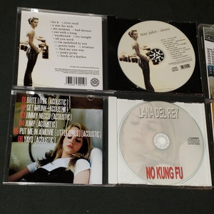 May include: A collection of six compact discs, including Lana Del Rey's "No Kung Fu" album, and a CD with the text "May Jailer - Sirens". The CDs are in jewel cases with white and black backgrounds. The back of one CD case lists the track titles for "Brite Lites (Acoustic)" by Lana Del Rey.