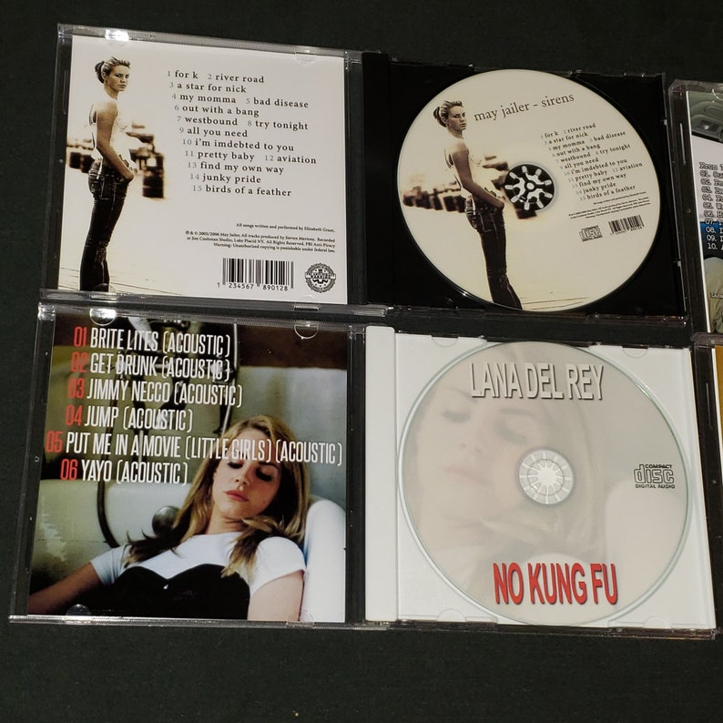 May include: A collection of six compact discs, including Lana Del Rey's "No Kung Fu" album, and a CD with the text "May Jailer - Sirens". The CDs are in jewel cases with white and black backgrounds. The back of one CD case lists the track titles for "Brite Lites (Acoustic)" by Lana Del Rey.