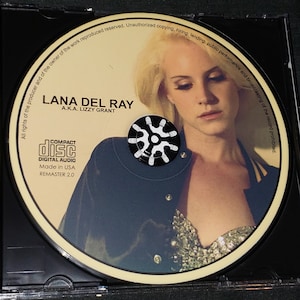 Puede incluir: Un CD con un retrato de Lana Del Ray, con el texto "LANA DEL RAY A.K.A. LIZZY GRANT" y "COMPACT DISC DIGITAL AUDIO Made in USA REMASTER 2.0".