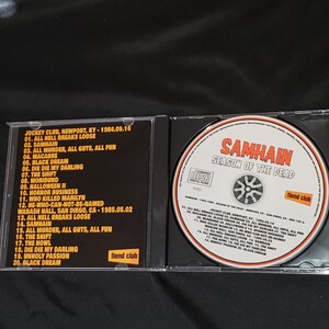 May include: A black CD case with a black and white image of a skull on the front. The case is labeled "Samhain Season of the Dead" and features the band's name in white text. The back of the case lists the tracklist for the album, which includes songs like "All Hell Breaks Loose", "Die Die My Darling", and "Unholy Passion".