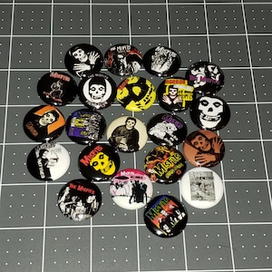 May include: A collection of 18 round Misfits band pins. The pins feature various band logos, album covers, and images of the band members. The pins are black, white, red, yellow, and blue.