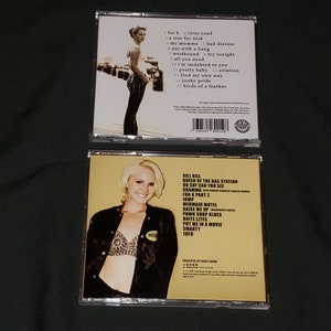 Puede incluir: Dos estuches de CD con fondo amarillo. La parte delantera del estuche presenta a una mujer con cabello rubio que lleva una blusa negra y un sujetador blanco. La parte posterior del estuche enumera los t&iacute;tulos de las canciones "Kill Kill", "Queen of the Gas Station", "On Say Can You See", "Gramma", "For K Part 2", "Jump", "Mermaid Motel", "Raise Me Up", "Pawn Shop Blues", "Brite Lites", "Put Me in a Movie", "Smarty" y "Yayo".
