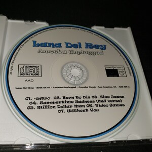 K&ouml;nnte beinhalten: Eine Compact Disc mit einem blauen und wei&szlig;en Rand. Die Disc hat ein wei&szlig;es Label mit dem Text "Lana Del Rey Amoeba Unplugged" in Schwarz. Die Trackliste ist unter dem Titel gedruckt: "01. -intro- 02. Born To Die 03. Blue Jeans 04. Summertime Sadness (2nd verse) 05. Million Dollar Man 06. Video Games 07. Without You".
