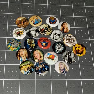 May include: A collection of 16 round buttons featuring images of the Allman Brothers Band. The buttons feature various band members, album covers, and band logos. The buttons are a mix of colors, including red, blue, green, yellow, and white.