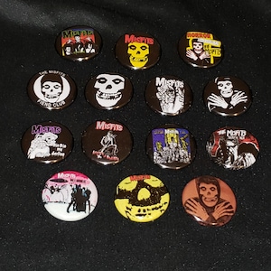 May include: A collection of 12 Misfits band pinback buttons. The buttons feature various album covers, band logos, and images of the band members. The buttons are black, white, red, yellow, and blue.