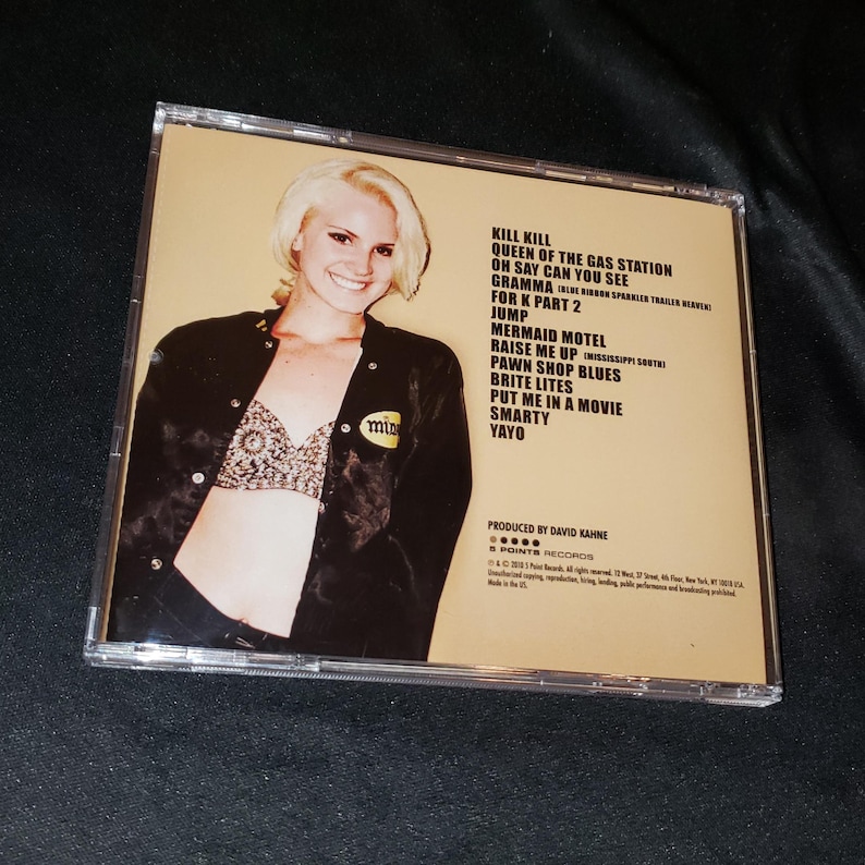 May include: A CD case with a yellow background and a woman wearing a black jacket and a sparkly bra. The text on the back of the case reads "Kill Kill, Queen of the Gas Station, On Say Can You See, Gramma, For K Part 2, Jump, Mermaid Motel, Raise Me Up, Pawn Shop Blues, Brite Lites, Put Me In A Movie, Smarty, Yayo".