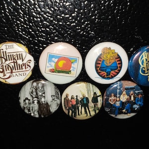 May include: Seven round buttons with different images of the Allman Brothers Band. The buttons feature a peach, a mushroom, a band photo, and the band's logo in different colors and styles.