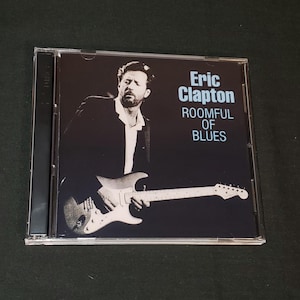 May include: A CD case with a black background featuring Eric Clapton playing a white electric guitar. The text on the case reads "Eric Clapton Roomful of Blues".